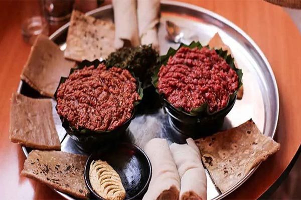 A traditional Ethiopian food kitfo with two bowls of red and brown curries, injera rolls, kocho, and koba on a metal tray.
