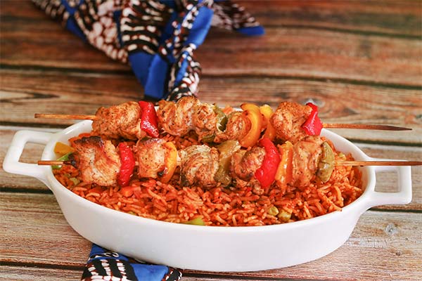 A white dish filled with vibrant jollof rice topped with skewered, grilled chicken and colorful bell peppers.