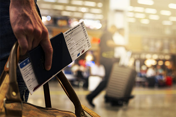 Close-up of a person holding a boarding pass and passport in an airport. The background shows travelers with luggage.